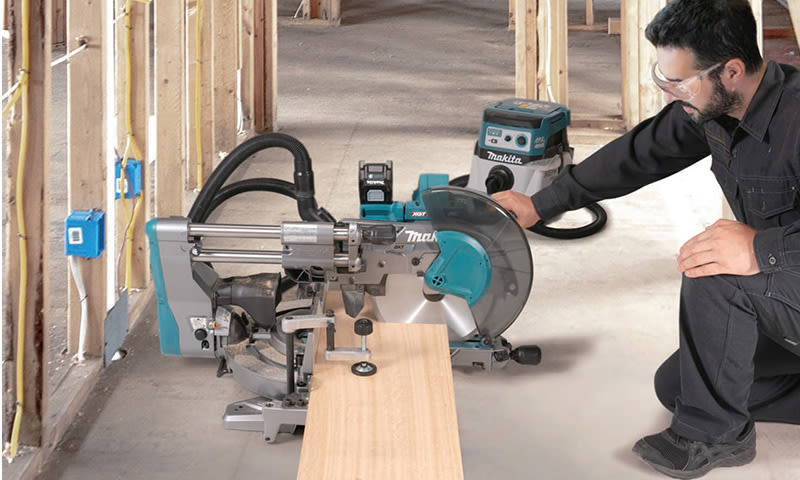 Arbeider ved teal/grå gjæringssag i uferdig bygg skjærer plankebit.