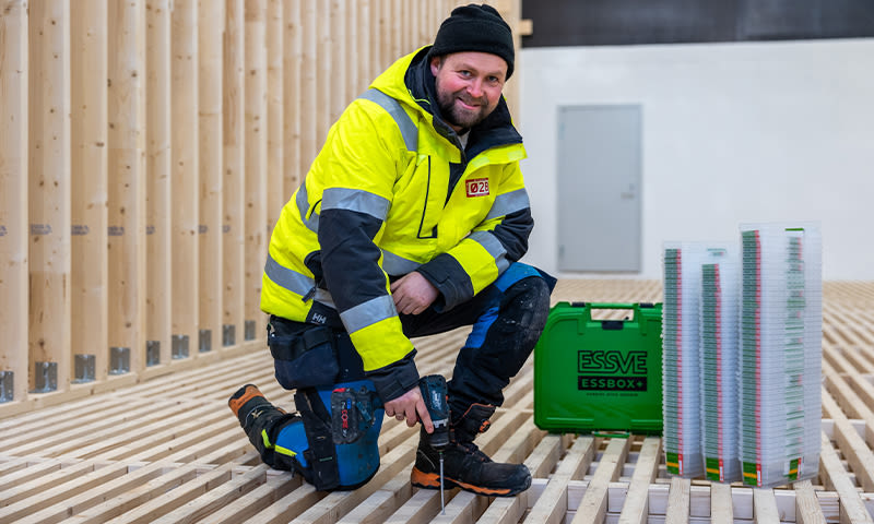 Anleggsarbeider i gul refleksjakke med drill på trebjelkelag, ved grønn ESSVE-kasse og skruer