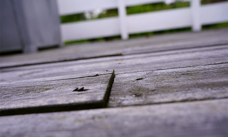 Nærbilde av slitte, grå terrassebord med hvitt gjerde og grønt gress i bakgrunnen
