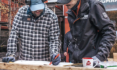 To håndverkere i rutete skjorte og svart jakke måler en treplanke ved byggeplass med sveisesprut.