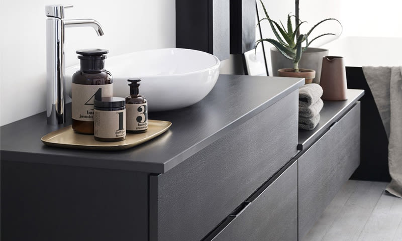 Moderne bad med mørk grå vaskeseksjon, hvit bolleservant, kromkran, brune flasker og sukkulent.