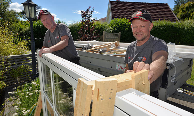 To håndverkere i grå t-skjorter og rødsvarte capser bærer en hvit vindusramme på tilhenger i hage.
