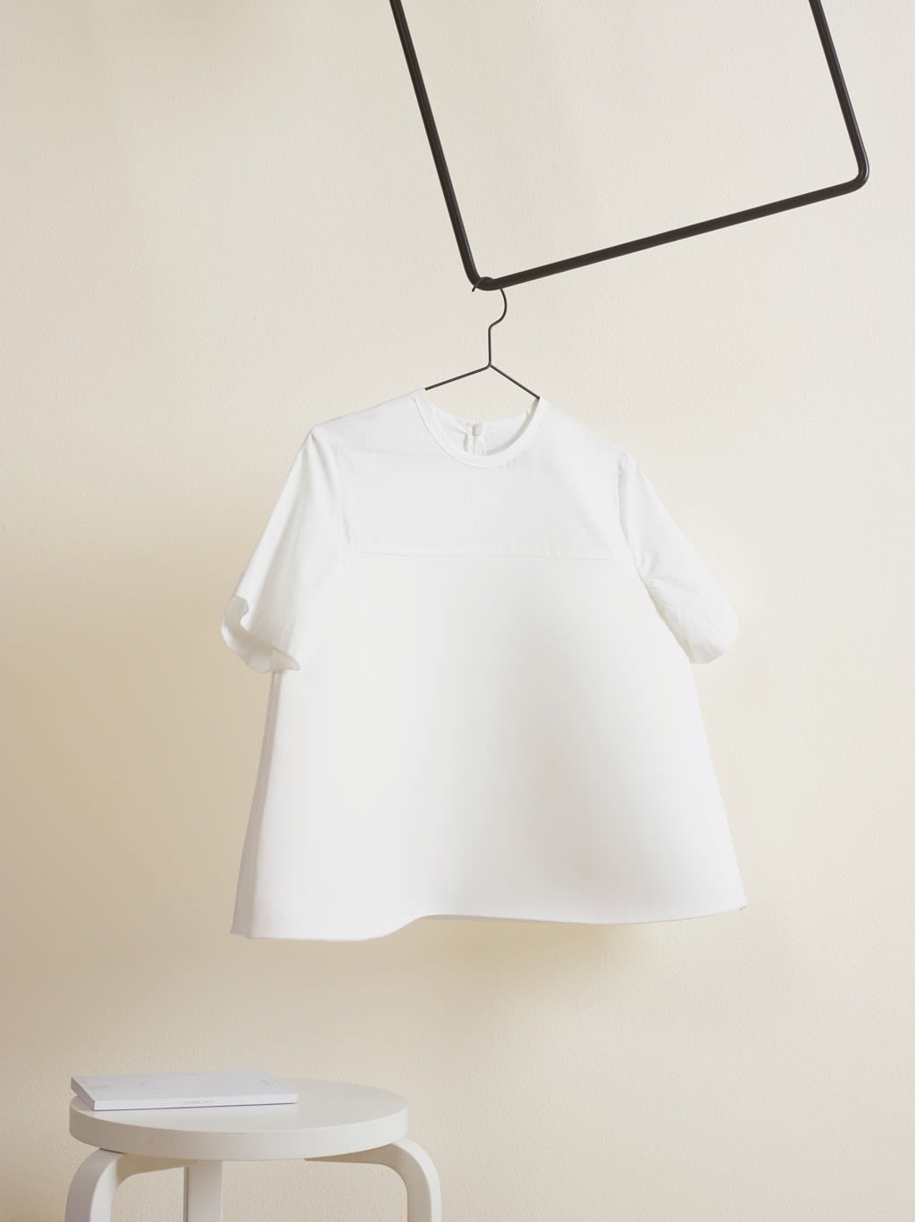 Hvit vid t-skjorte henger i svart metallramme mot kremfarget vegg, hvit krakk med bok