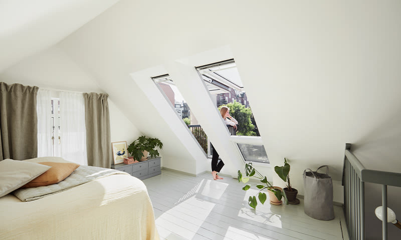 Lyst loftsoverom med skråtak, hvite vegger, store takvinduer, grønne planter og kvinne ved vindu.
