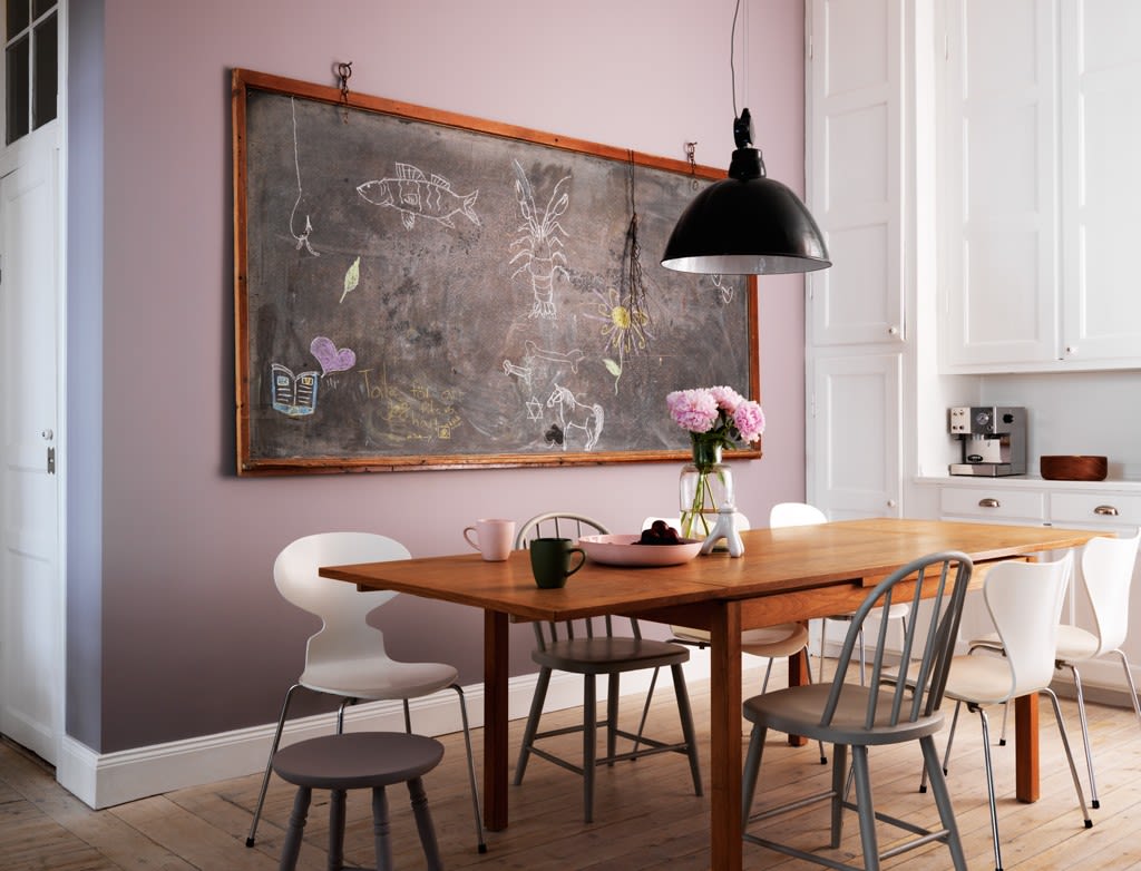 Treverksbord med vase med rosa blomster, ulike stoler, svart hengelampe, rosa vegg og svart tavle.