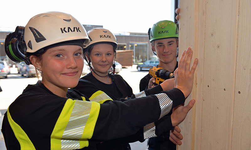 Tre arbeidere med KASK-hjelmer og gule refleksvester skrur fast lyst trepanel ute.