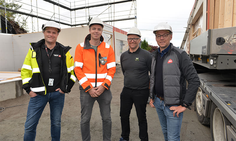 Fire byggarbeidere med vernehjelmer og refleksjakker foran stillas og lastebil med elementer.