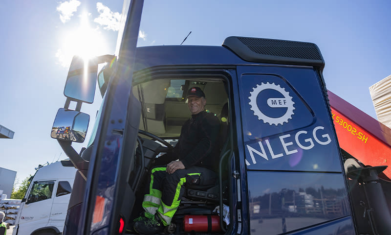 Fører i gul-grønn refleksbukse i blå MEF Anlegg-lastebil under solskinn, rød kran i bakgrunnen