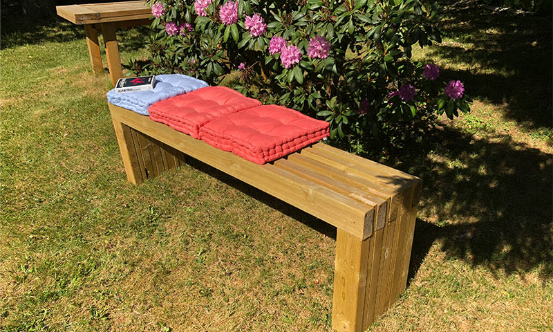 Trebenk under rosa rhododendron i sol, med lyseblått og rødt sitteunderlag og bok