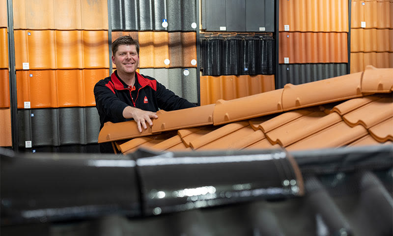 Montør holder oransje takstein foran vegg med sorte og terrakotta takstein.