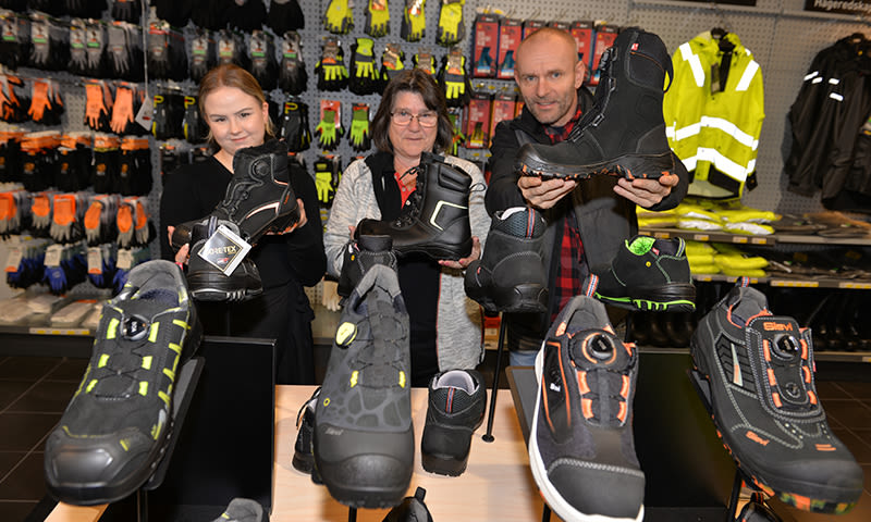 Tre personer viser svarte og oransje vernesko foran hylle med hansker og gule jakker.
