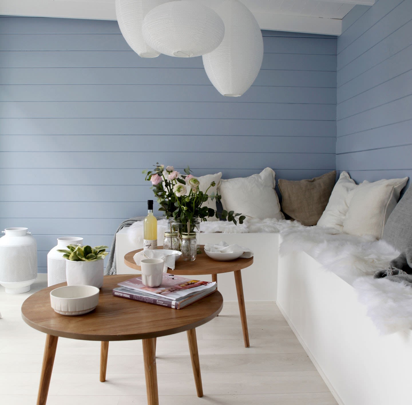 Lyst rom med lysblå panelvegg, hvit sittebenk med puter, trebord med blomster.