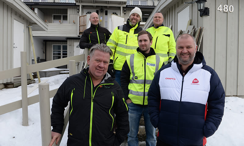 Byggearbeidere i gule og mørke jakker står foran snødekket grå hytte.