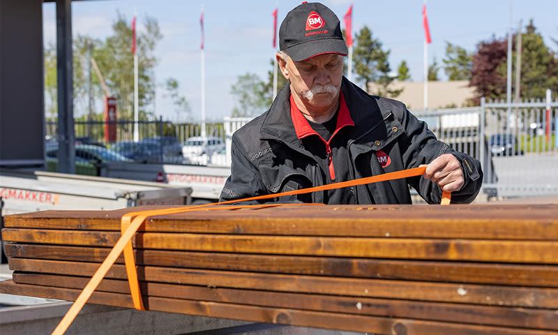 Ansatt strammer oransje stroppebånd rundt brune treplanker utenfor byggevarebutikk
