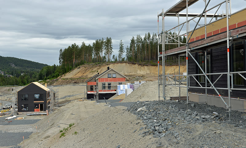 Flere uferdige svarte trehus med stillas på grå steinur ved furuskog under overskyet himmel.
