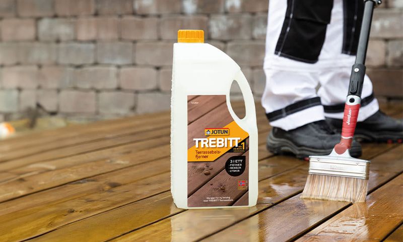Hvit Jotun Trebitt-kanne med oransje kork på vått brunt terrassegulv mens hånd med pensel renser