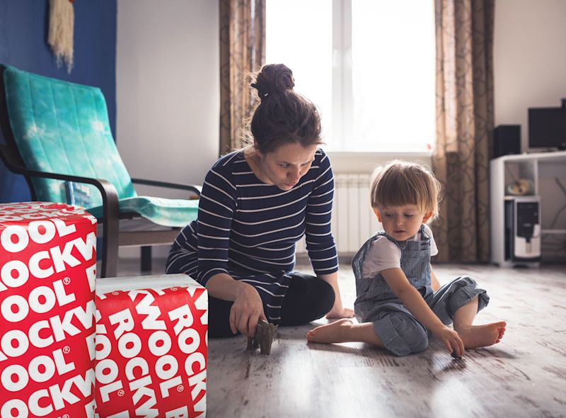 Mor i stripet topp og barn i denim overalls leker på lyst gulv ved røde ROCKWOOL-kasser.