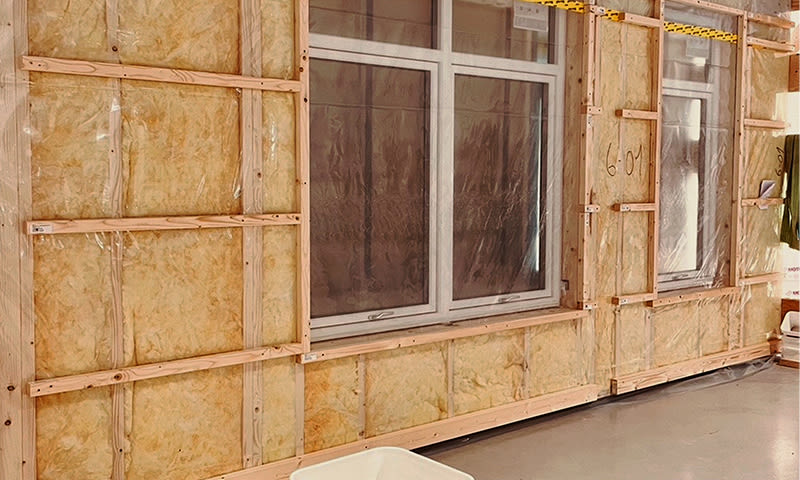 Innvendig vegg i oppussing med gul isolasjon, trelektverk og to plastdekte hvite vinduer