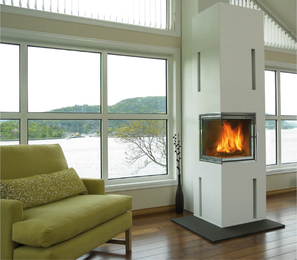 Moderne stue med grønn lenestol, hvit peis med flammer og panoramavindu mot vann og skog.