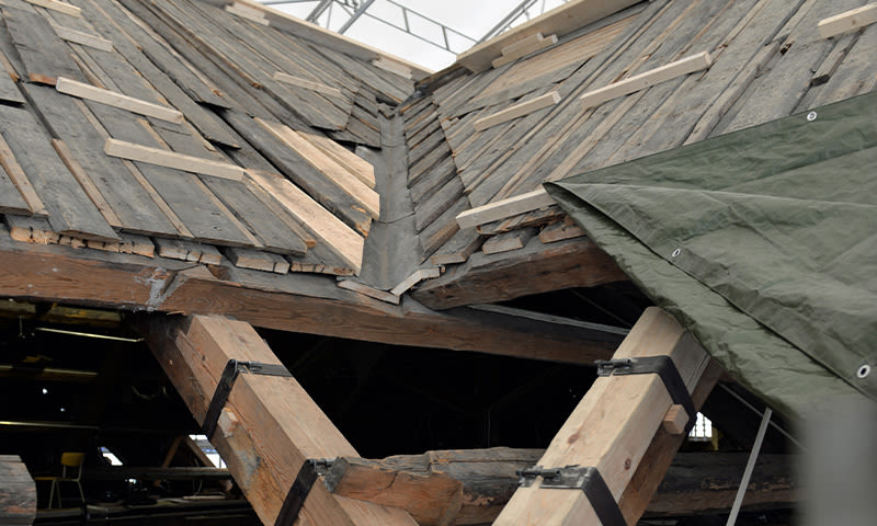 Delvis demontert tak av grå skiferplater, synlige trebjelker og grønn presenning under reparasjon