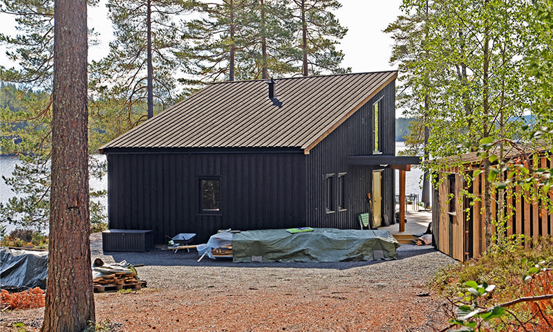 Svart moderne hytte i furuskog ved innsjø, grusplass med dekkede materialer