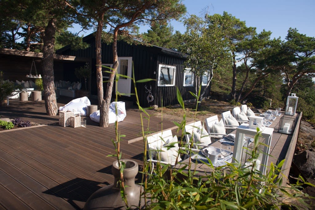 Lang treterrasse med dekket bord, hvite lanterner og puter ved svart hytte i furuskog