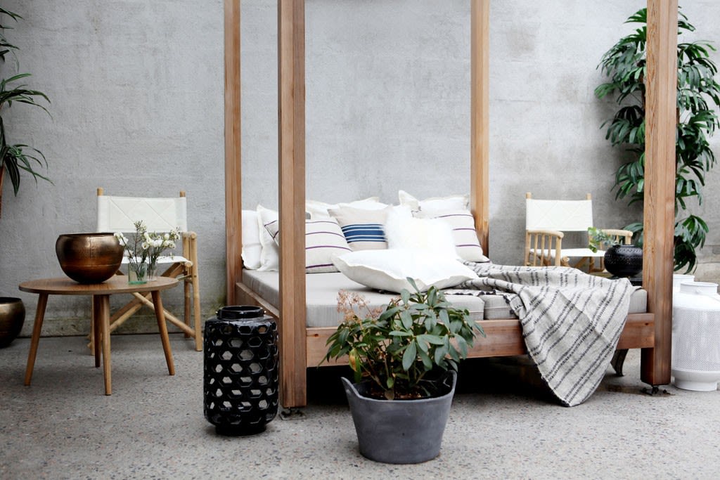 Ute-lounge med trebaldakin, hvite puter, stripete pledd, bambusstoler og grønne planter.