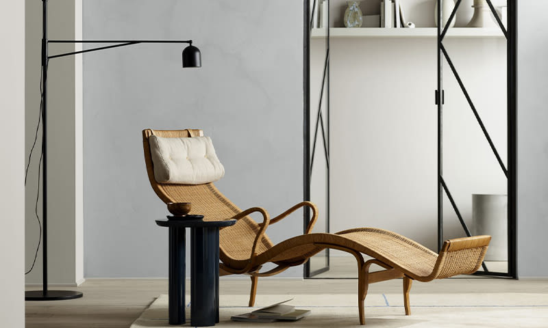 Lysegrå stue med kurvestol i rotting med hvit pute, fotstøtte, svart gulvlampe og metallhylle.