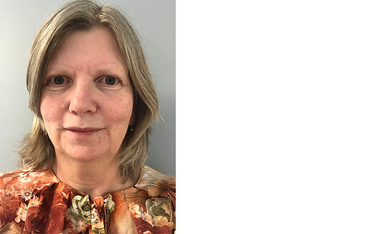 Kvinne med skulderlangt blondt hår i blomstrete oransje- og grønnfarget bluse foran lys grå vegg.