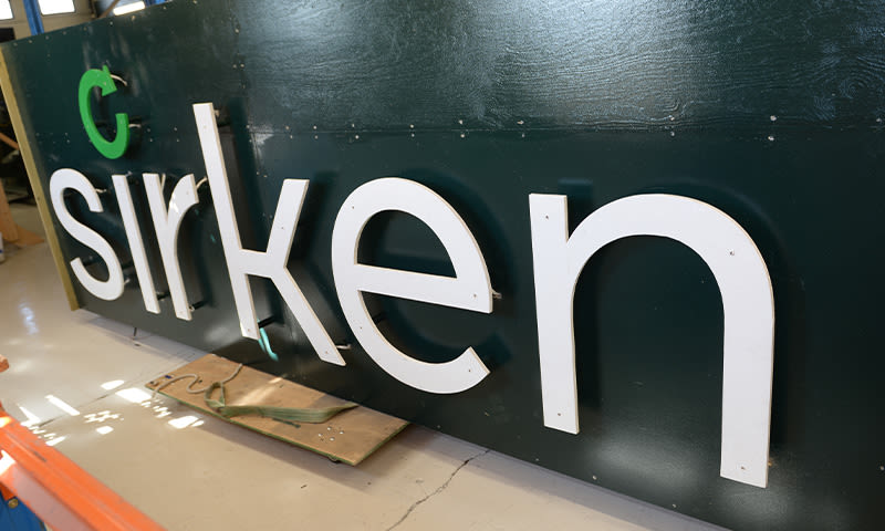 Hvite 3D-bokstaver sirkelen og grønn C på mørkegrønn skilt liggende på verkstedgulv.