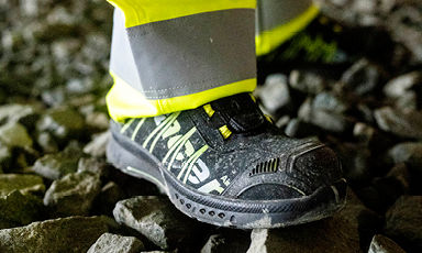 Nærbilde av svart-gule vernesko og gul refleksbukse på grus.
