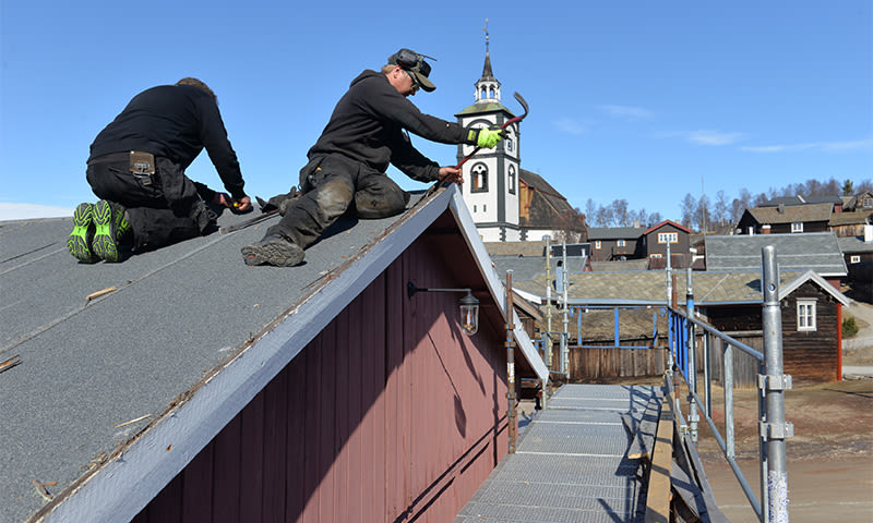 To taktekkere på rødt tretak under blå himmel, kirke og stillas i bakgrunnen.