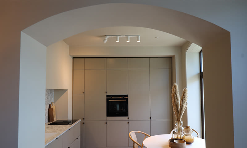 Moderne kjøkken i lyse gråtoner gjennom bueåpning, skap og ovn, hvitt bord og pampasgress