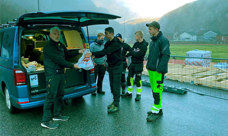 Fem håndverkere i arbeidsklær ved åpen blå varebil på byggeplass i fjordlandskap.