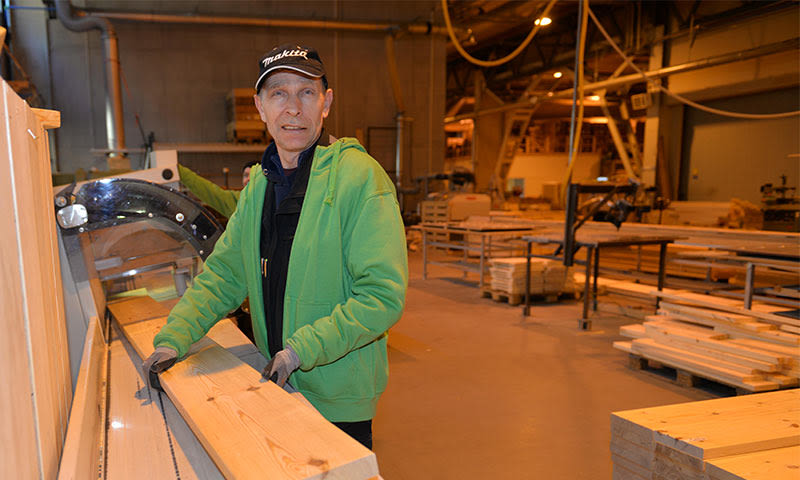 Arbeider i grønn hettegenser skjærer lys treplanke på sagmaskin i trevarefabrikk