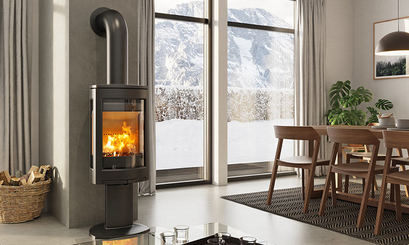 Svart vedovn med flammer ved vindu med utsikt til snødekt fjell, trebord og stoler.