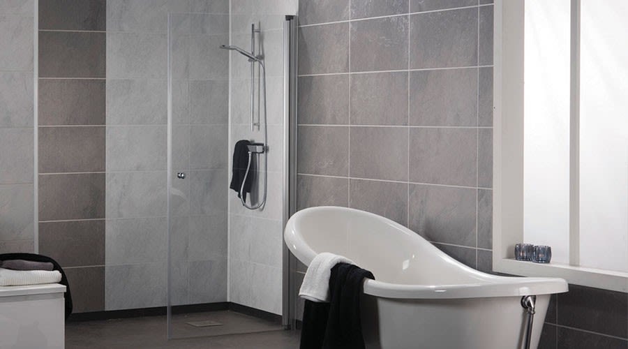 Moderne bad med hvitt frittstående badekar, grå veggfliser, glassdusj og sorte håndklær.