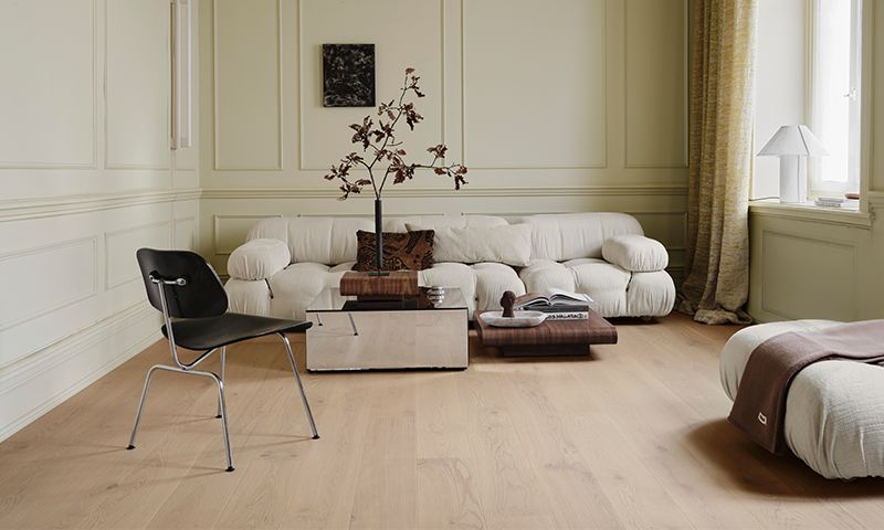 Lyst kremhvit stue med buet sofa, svart stol, lavt glassbord med grener, lyse tregulv