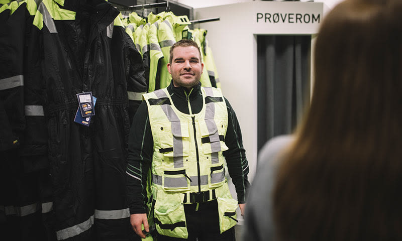Person i gulgrønn refleksvest foran rad med mørke arbeidsklær ved prøverom