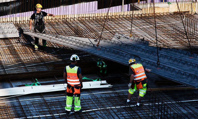 Tre anleggsarbeidere i oransje vester og gule hjelmer løfter betongbjelke over armeringsjern.