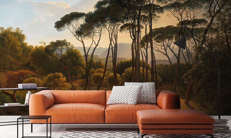Oransje skinnsofa med to mønstrede puter foran mural av grønne furutrær i dempet kveldlys.