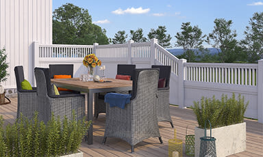 Uteplass på treterrasse med grå flettstoler rundt bord, solsikker, fargerike puter og hvitt gjerde.