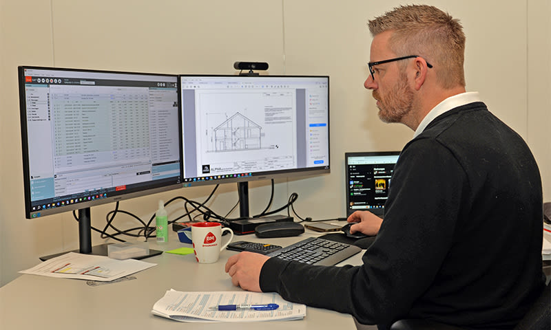 Mann i kontor med tre skjermer, hus-tegning på midtskjerm, rød kopp på pult.