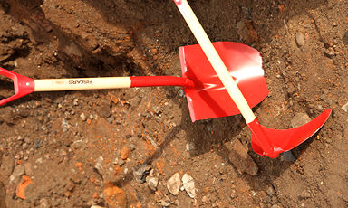 To røde hageredskaper (spade og hakke) med lyse treskaft på grusete jord.