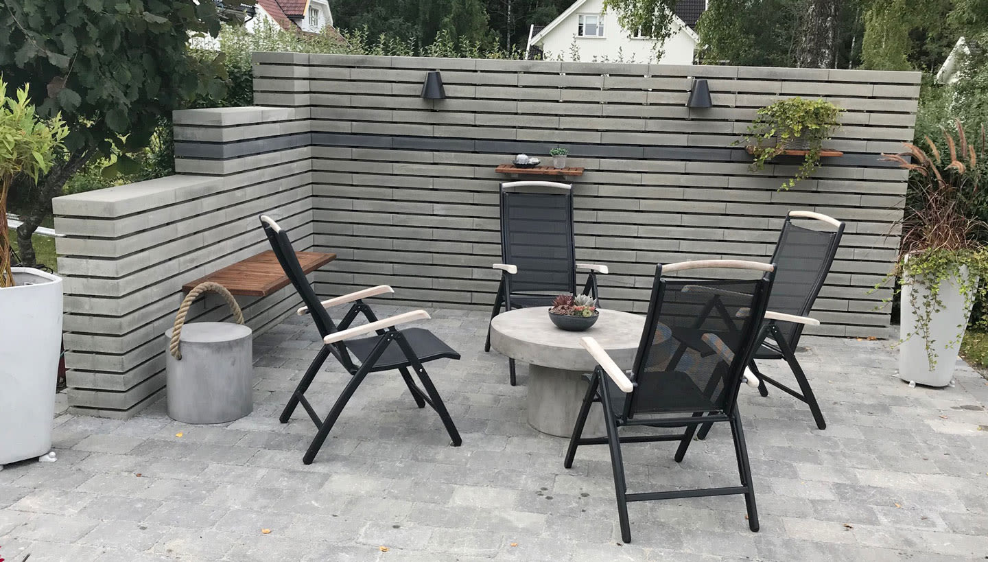 Moderne uteplass med grå slatvegg, fire svarte stoler rundt betongbord med sukkulenter.