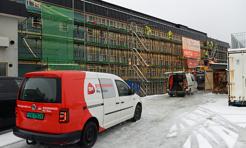 Rød-hvit Byggmakker varebil foran snødekt byggeplass med stillas, grønn presenning og arbeidere.