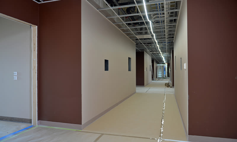 Lang korridor under oppussing med beige gulv, brune vegger, eksponert tak og lysrør