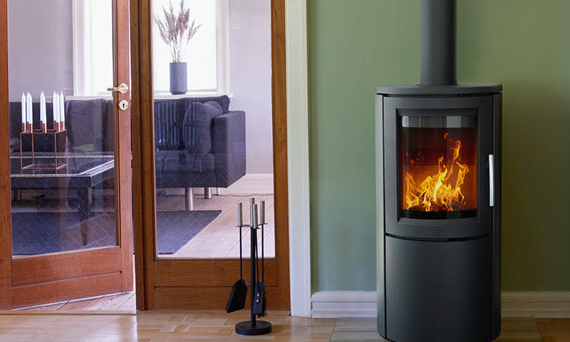 Svart vedovn med ild mot grønn vegg, peisverktøy og glassdør til stue med grå sofa