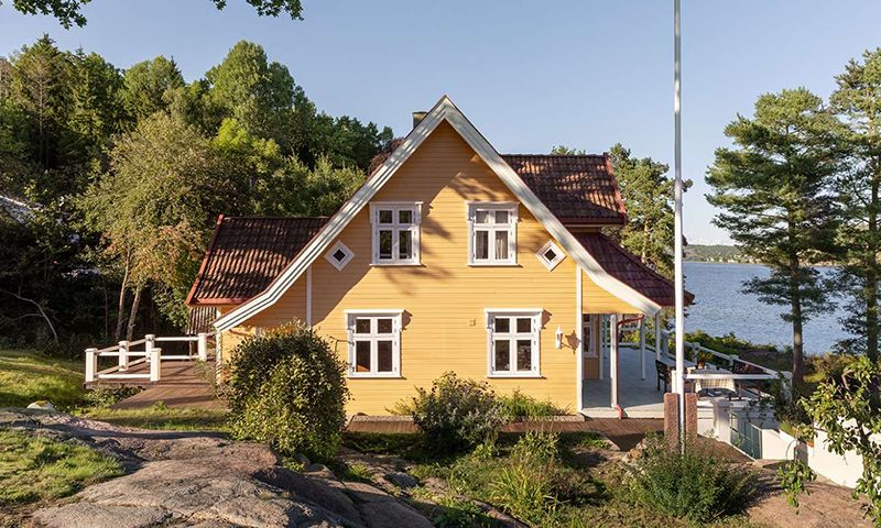 Gult trehus med rød takstein og hvite vinduer ved innsjøkant, omgitt av skog og svaberg.
