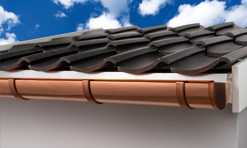 Nærbilde av kobberrenne under mørk takstein, mot blå himmel med hvite skyer.